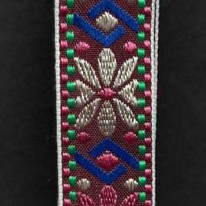 Close-up of embroidered ribbon with symmetrical floral pattern in metallic pink and silver threads bordered by green stitches.