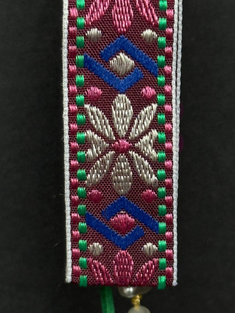 Close-up of embroidered ribbon with symmetrical floral pattern in metallic pink and silver threads bordered by green stitches.