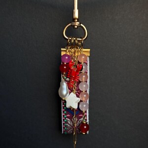 Op de afbeelding: Een decoratieve sleutelhanger met een gouden sluiting en een basis van stof met patronen. Het heeft een rode kreeftbedel, diverse kralen in roze, rood en wit, en een witte sterbedel. De sleutelhanger hangt tegen een donkere achtergrond.