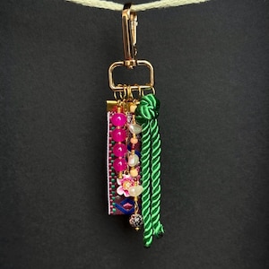 Op de afbeelding: Een decoratieve sleutelhanger met een goudkleurige sluiting en een rechthoekige stoffen bedel. De bedel is versierd met roze kralen, een bloemaccent en een groene kwast. De sleutelhanger hangt tegen een donkere achtergrond.
