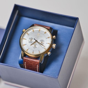 Reloj de pulsera elegante para hombre, estilo cronógrafo en tono dorado con correa de piel marrón.