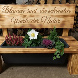 Könnte beinhalten: Holzbank mit einem Pflanzkasten gefüllt mit Blumen und Grün. Die Bank hat eine Rückenlehne mit dem deutschen Text "Blumen sind die schönsten Worte der Natur" eingraviert. Die Bank ist aus hellbraunem Holz.