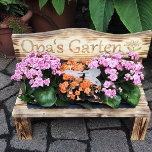 Könnte beinhalten: Eine kleine Holzbank mit Flammoptik, die den Schriftzug "Opa's Garten" und eine Blumengravur trägt. Auf der Bank stehen drei getopfte Kalanchoe-Pflanzen mit leuchtend rosa und orangefarbenen Blüten sowie dunkelgrünen Blättern in einer Reihe.