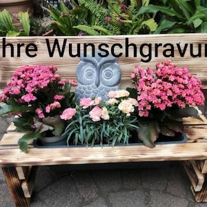 May include: Wooden bench planter with pink flowers and a gray owl statue. The bench has a distressed finish and the text "Ihre Wunschgravur" is visible on the back. A decorative piece for the garden.