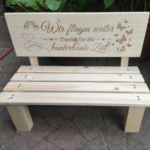Könnte beinhalten: Eine helle Holzbank mit einer gravierten Rückenlehne, die den deutschen Text Wir fliegen weiter Danke für die kunterbunte Zeit sowie eingravierte Abbildungen eines Regenbogens, eines Herzens, von Wolken und Schmetterlingen zeigt.