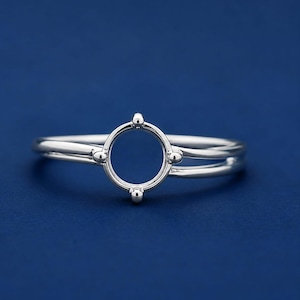May include: A polished silver ring featuring a circular setting at the center. The band is split into two thin, parallel lines that converge at the sides. The circular setting is accented with four small, evenly spaced spherical prongs.