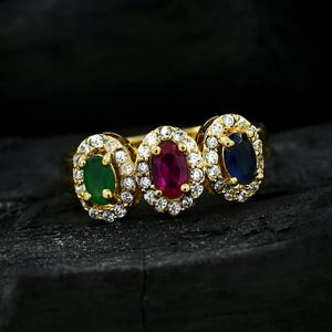 May include: A gold ring featuring three oval-cut gemstones in a row: a green stone, a red stone, and a blue stone. Each gemstone is surrounded by a halo of small, clear, round-cut white stones set in a prong setting on a gold band.
