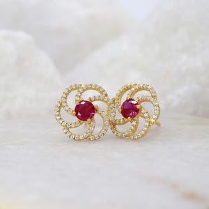 Puede incluir: Un par de pendientes de botón dorados con una piedra preciosa roja de corte redondo en el centro, rodeada por un marco floral en espiral incrustado con pequeñas piedras blancas en pavé. El diseño crea un patrón simétrico con un acabado metálico.