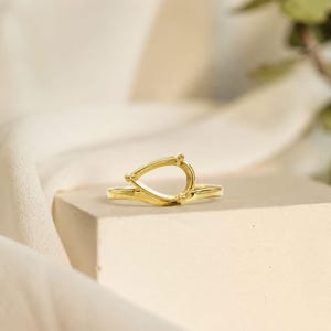 May include: A gold ring featuring a teardrop-shaped setting at the top. The band is smooth and polished, with a delicate, open-frame design that forms a pointed oval shape. The metal has a bright, reflective finish throughout the entire piece.
