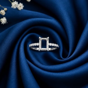 May include: A silver-toned ring setting featuring a rectangular prong head designed to hold a center stone. The band is accented with a row of small, clear, round-cut stones embedded along each side of the shank, creating a polished appearance.