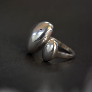 May include: A silver ring with a unique design. The ring features two oval-shaped, polished silver elements, one larger than the other, connected by a band. The ring is set against a dark background, highlighting its reflective surface.