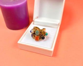 Könnte beinhalten: Ein goldfarbener Ring mit mehreren bunten Edelsteinen, präsentiert in einer weißen Schmuckschatulle. Der Ring hat Steine in Blau, Grün, Orange und Schwarz. Eine violette Kerze befindet sich links von der Schatulle auf einer pfirsichfarbenen Oberfläche.