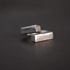 Könnte beinhalten: Ein silberfarbener Bypass-Ring mit zwei rechteckigen Stabenden. Das Metall hat eine glatte, polierte Oberfläche mit sichtbaren Lichtreflexionen entlang der Kanten der Stäbe. Der Ring ist offen gestaltet, wobei sich die beiden rechteckigen Segmente leicht überlappen.