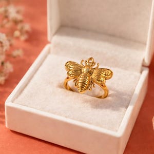 May include: A gold-colored bee ring displayed in a white jewelry box. The ring features a detailed bee design with textured wings and body. The background is a soft peach color with a hint of flowers.