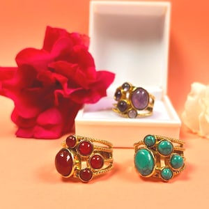 Peut inclure: Trois bagues dorées ornées de pierres précieuses colorées. Une bague présente une grande pierre rouge ovale entourée de petites pierres rouges. Une autre bague a une grande pierre turquoise ovale avec de petites pierres turquoises.