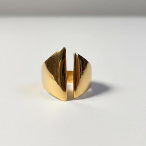 May include: A gold ring with a geometric design. The ring has two triangular shapes joined at the base, with a vertical split in the middle. The ring is made of a shiny, reflective metal.
