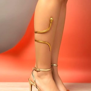 Könnte beinhalten: Goldfarbene Schlangen-Beinmanschette und passende High-Heel-Sandalen. Die Schlangenmanschette umschlingt das Bein, der Kopf der Schlange befindet sich in der Nähe des Knies. Die Sandalen haben einen dünnen Knöchelriemen und einen Stiletto-Absatz.