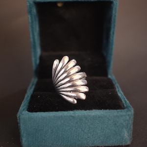 May include: A silver ring with a fan-shaped design, presented in an open teal velvet jewellery box with a black interior. The ring's design features multiple curved, parallel lines. The box is on a dark surface.