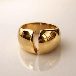 May include: A gold cuff bracelet with a split design. The polished surface reflects light, creating highlights and shadows. The bracelet's rounded shape and smooth finish give it a modern, minimalist aesthetic. This piece of jewellery is suitable for everyday wear.