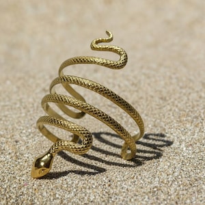 Può includere: Un bracciale dorato a forma di serpente, avvolto su se stesso. La testa del serpente è in basso e la coda in alto. Il bracciale è appoggiato su una superficie sabbiosa.
