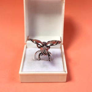May include: A silver-toned metal ring shaped like a fly with detailed wings and legs, positioned inside a white rectangular jewelry box. The ring features a metallic finish with visible textures on the insect's body and wings, resting on a white base.