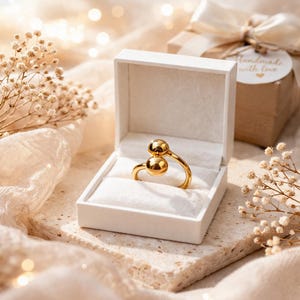 Op de afbeelding: Een goudkleurige ring met twee bolvormige accenten, gepresenteerd in een open witte doos met een zacht interieur. De ring ligt op een getextureerd, lichtgekleurd oppervlak, met een geschenkdoos en decoratieve elementen op de achtergrond.