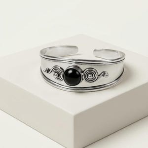 May include: A silver-toned cuff bracelet featuring a polished black circular stone at the centre. The metal band is decorated with symmetrical swirling scrollwork patterns on both sides of the stone, creating a detailed and structured design.