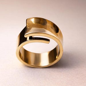 May include: A gold-colored ring with a unique, modern design. The ring features a thick band with a curved, overlapping element. The polished surface reflects light, highlighting the ring's smooth texture and shape. A stylish piece of jewelry.