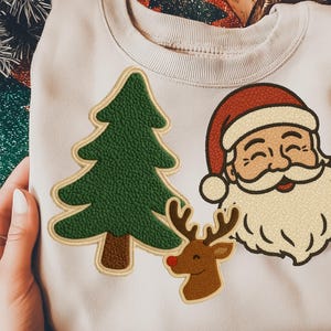 Puede incluir: Una sudadera beige con un diseño de árbol de Navidad, Papá Noel y reno. El árbol es verde, Papá Noel tiene un gorro rojo y barba blanca, y el reno tiene una nariz roja. La sudadera es sostenida por manos.