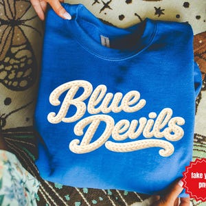 May include: Royal blue sweatshirt with the words "Blue Devils" in a cream-colored, textured font. The sweatshirt is folded and held by hands, resting on a patterned surface.