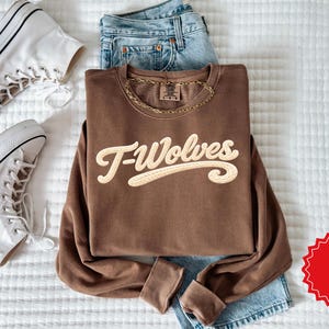 May include: A brown sweatshirt with the text "T-Wolves" in a cream-colored, stylized font. The sweatshirt is paired with light-wash denim jeans, white high-top sneakers, and a gold chain necklace. The items are arranged on a white, textured surface.