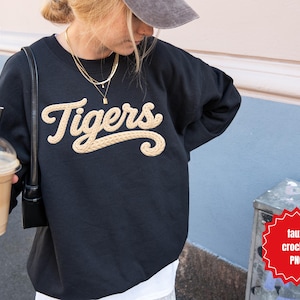 May include: Black sweatshirt with the word "Tigers" in a cream-colored, textured font. The sweatshirt is worn by a person holding a beverage in a clear plastic cup with a black straw.