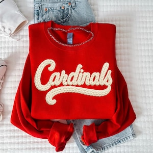 May include: A red sweatshirt with the word "Cardinals" in a cream-colored, textured font. The sweatshirt is paired with blue jeans and white high-top sneakers. A silver chain necklace is draped on the sweatshirt.