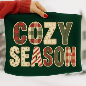May include: A dark green pillow with the words "COZY SEASON" in a patchwork design. The letters are made of various fabrics, including plaid, floral, and herringbone patterns. The pillow is held by a person with red nail polish.