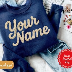 May include: Navy blue sweatshirt with "Your Name" in a cream-colored, faux crochet design. The word "Personalized" is on a small orange rectangle. The image includes a pair of blue jeans and a notebook.