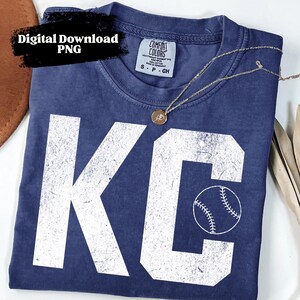 以下が含まれることがあります： ネイビーブルーの半袖Tシャツで、白いかすれた文字でKCとプリントされ、Cの文字の中に野球ボールのグラフィックが描かれています。首元にはComfort Colorsのラベルがあり、黒いテキストでDigital Download PNGと書かれています。