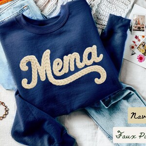May include: Navy blue sweatshirt with the word "Mema" in a cream-colored, braided font. The sweatshirt is displayed with blue jeans and a bracelet. The text "Navy" and "Faux Patches" are also visible.