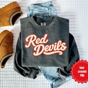 May include: A dark gray sweatshirt with the text "Red Devils" in a cream-colored, retro font with a red outline. The sweatshirt is paired with light-wash denim jeans and brown platform clogs. A red starburst graphic reads "faux crochet PNG".