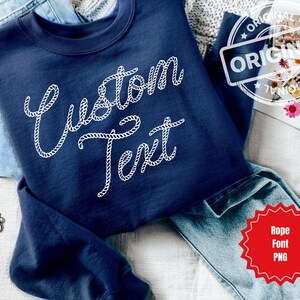 May include: Navy blue sweatshirt with the words "Custom Text" in a white rope font. The sweatshirt is folded on top of a pair of blue jeans. A gold chain bracelet is visible on the left side of the image.