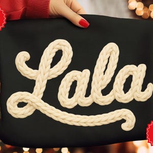 May include: Black tote bag with the word "Lala" in cream-colored, textured, rope-like letters. The bag is held by a person with red nail polish. The words "Faux Crochet" are in a red circle.