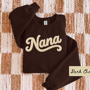 May include: A dark chocolate-coloured sweatshirt featuring the word "Nana" in a cream-coloured, textured script. The garment is displayed on a woven, neutral-toned surface. The words "Dark Chocolate" are in a cream-coloured box.