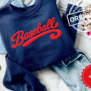 Pode incluir: Camisola azul marinho com a palavra "Baseball" em vermelho, com letras de crochê. A camisola está dobrada sobre um par de calças de ganga azuis. Uma pulseira de corrente dourada é visível no canto inferior esquerdo.