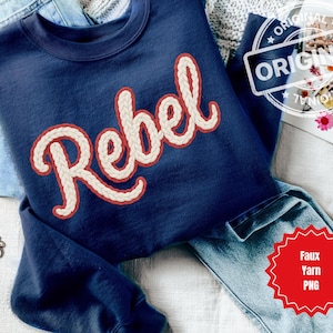 May include: Navy blue sweatshirt with the word "Rebel" in a cream-colored, braided font with a red outline. The sweatshirt is folded on top of blue jeans. A gold chain bracelet is visible.