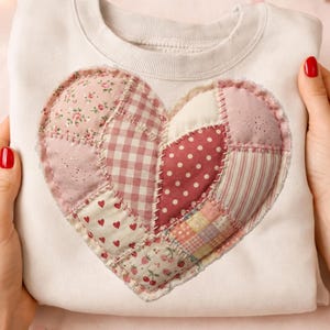 Op de afbeelding: Een witte sweatshirt met een groot patchwork hartontwerp. Het hart is gemaakt van verschillende roze en rood gekleurde stoffen, waaronder bloemen, stippen en ruitjes. De sweatshirt wordt vastgehouden door handen met rode nagellak.