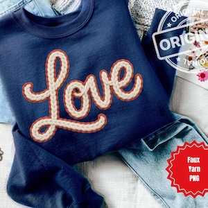 Può includere: Felpa blu navy con la scritta "Love" in un carattere testurizzato color crema con un contorno rosso. La felpa è piegata sopra dei jeans blu. È visibile un braccialetto a catena dorata.