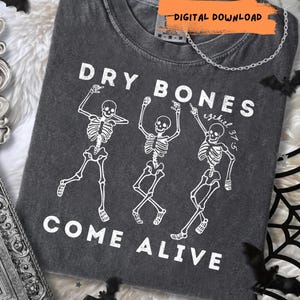 Puede incluir: Una camiseta gris carbón con un gráfico blanco de tres esqueletos bailando. El texto "DRY BONES" aparece sobre los esqueletos y "COME ALIVE" debajo, con "Ezekiel 37:5" escrito en letra cursiva junto al esqueleto de la derecha.