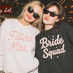 May include: Two women wearing sunglasses pose. One wears a white sweatshirt with "Future Mrs." in pink script and pearl accents. The other wears a black sweatshirt with "Bride Squad" in pink script and pearl accents. The image has a blurred background.