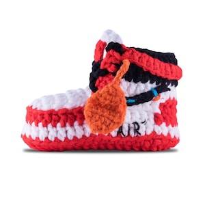 May include: A handmade crochet baby bootie in the style of a high-top sneaker. The bootie features red, white, and black yarn, with an orange tag and the word "AIR" on the side. The bootie is designed to resemble a popular athletic shoe.