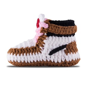 May include: A handmade crochet sneaker in brown, white, black, pink, and red. The shoe features a high-top design with a black stripe and a brown sole. The crochet stitches are visible, giving it a textured appearance.