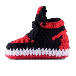 May include: A handmade crochet sneaker in a high-top style. The shoe features a color scheme of black, red, and white, with the upper predominantly black and red, and the sole and trim in white. The sneaker is designed to resemble a popular athletic shoe.